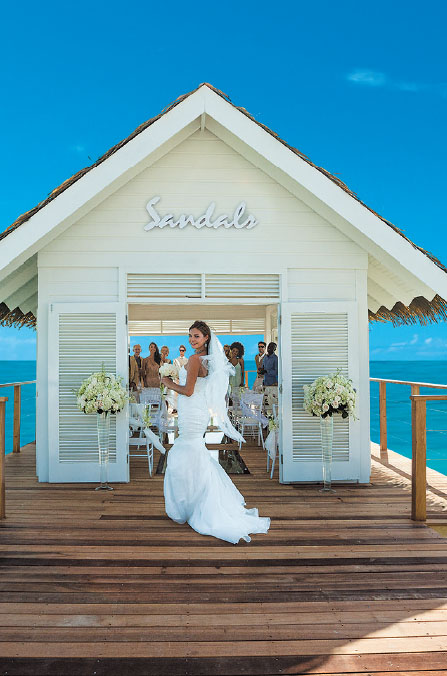 A bride stands in front of a white chapel on a dock leading out to a clear blue ocean and cloudless sky. The bride is smiling over her shoulder, bouquet in hand, and dress train flowing behind her on the dock. The bride is pictured at Sandals South Coast.