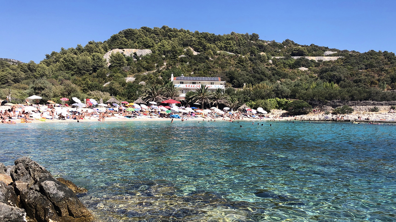 A beach in Hvar, Croatia is covered in white sandy beaches, colourful umbrellas and sunbathers on a cloudless afternoon. The shallow blue water is crystal clear.