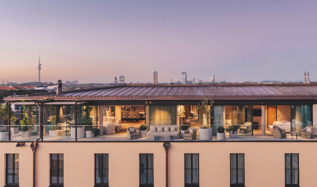 The rooftop terrace at Hotel Bayerischer Hof is decorated with several sets of warm furnishings and lots greenery in stone planters. The Frauenkirche and Olympiaturm are visible in the skyline in the background.