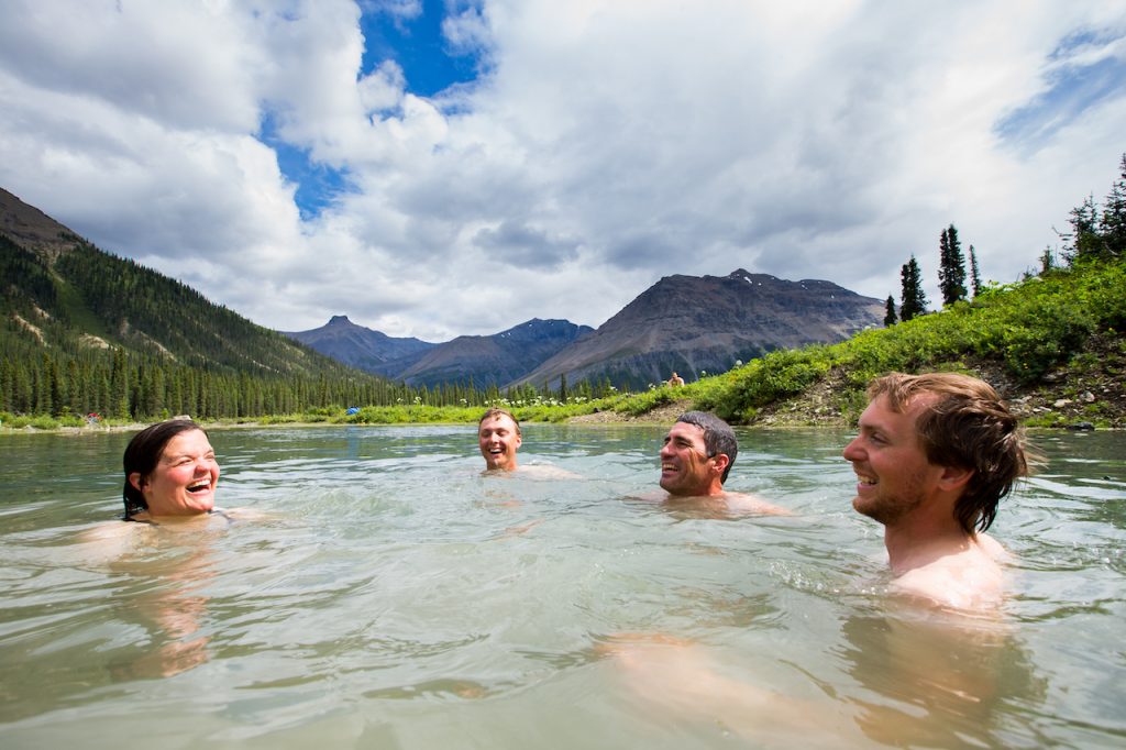 Four people swim in a lake with pine forests and mountains in the background. The four people are all smiling and laughing.