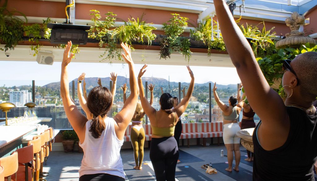 Over half a dozen people stand facing away from the camera, overlooking a city skyline, all in a raised arms yoga pose. There are ferns and other greenery decorated along the rooftop.