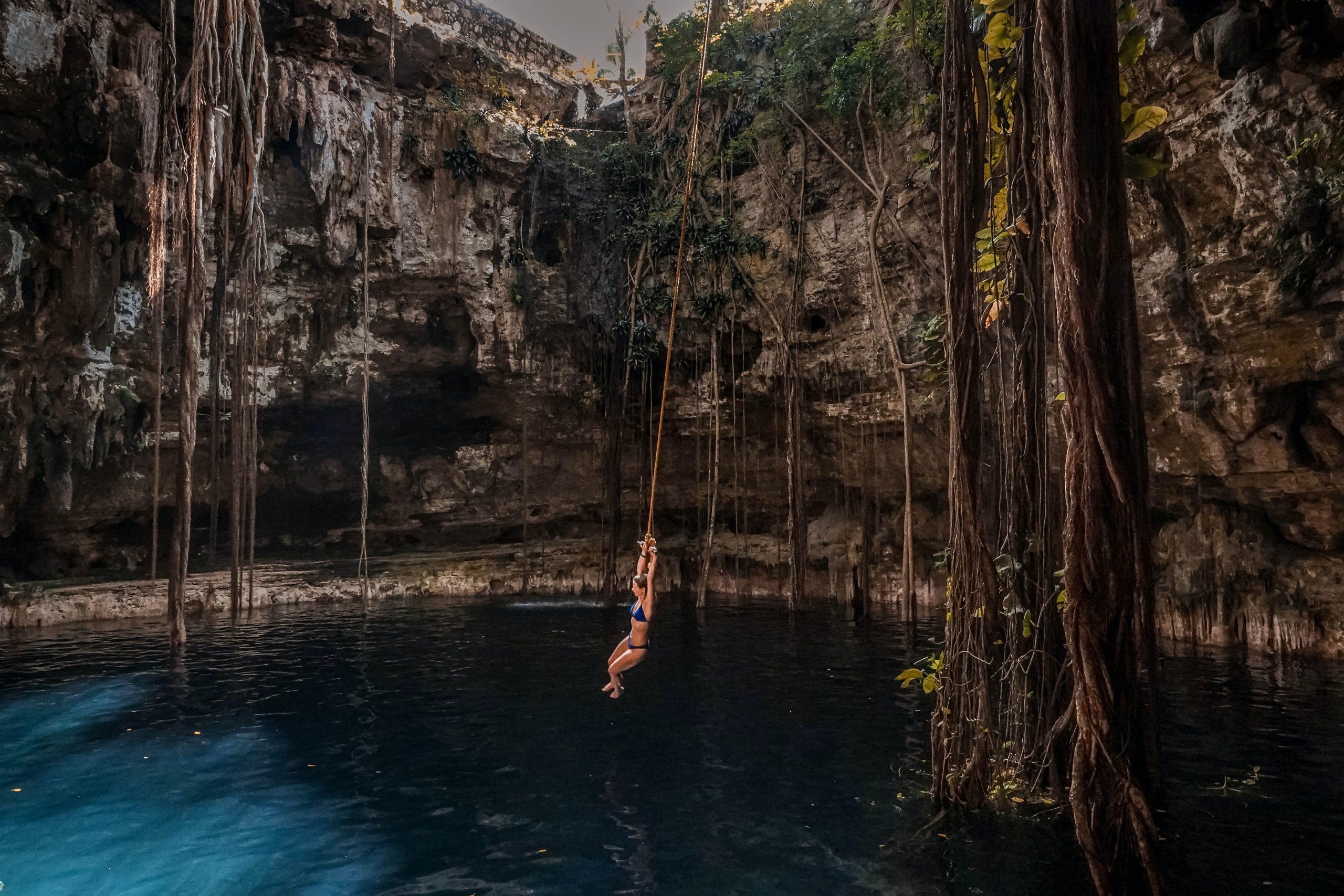 A person swings from a rope above a sunken lake or quarry. The clear pool of water is surrounded by rock walls and climbing roots and vines.